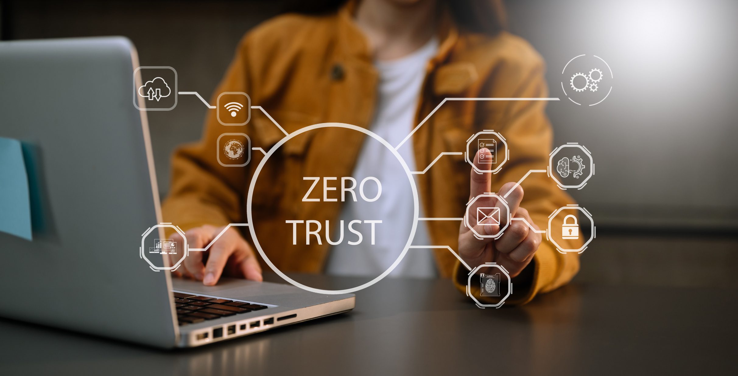 A woman in a yellow jacket using her laptop with a digital graphic depicting 'Zero Trust' in the forefront.