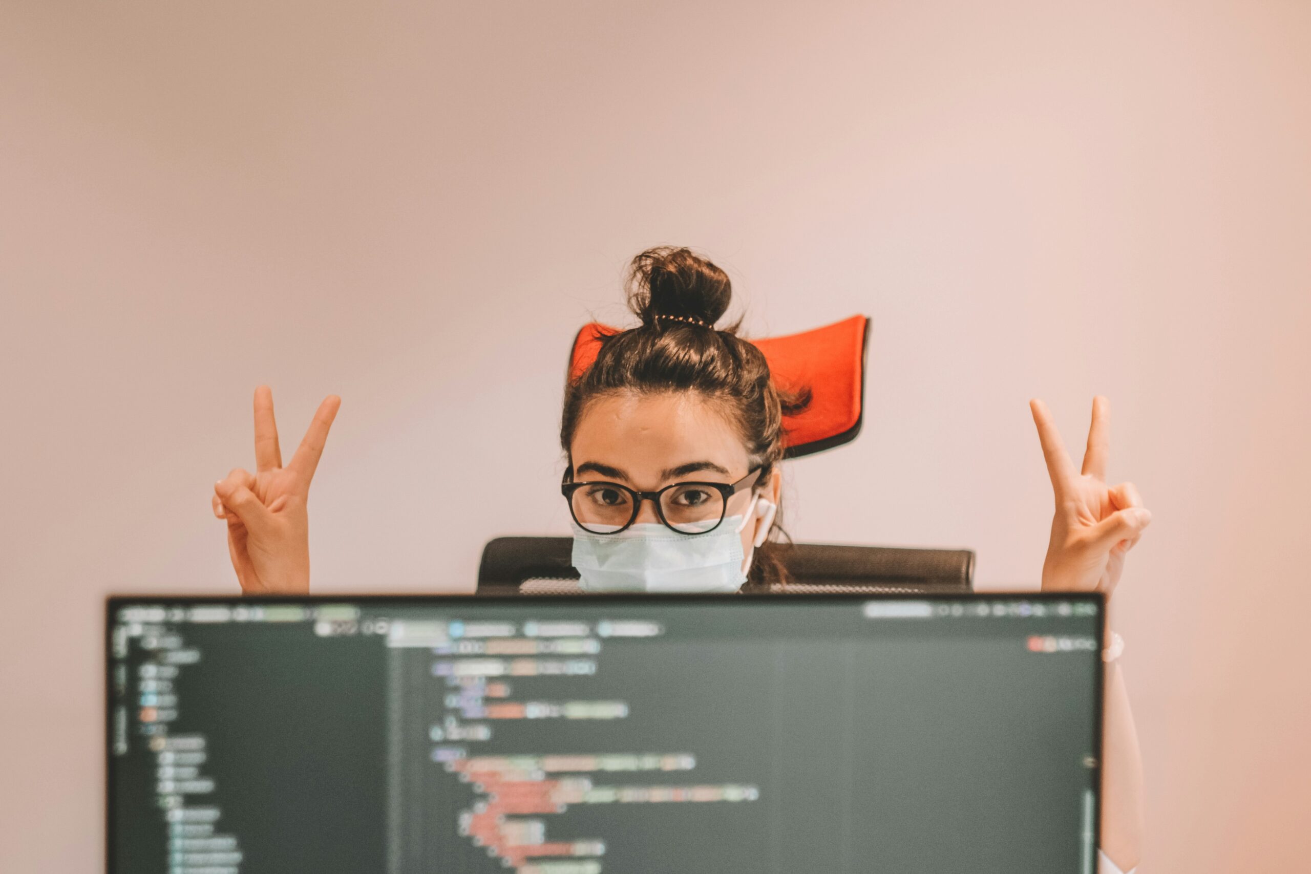 A woman wearing a protective mask and holding up both hands to make the 'peace' sign with her fingers can be seen sitting behind a computer screen showing code.