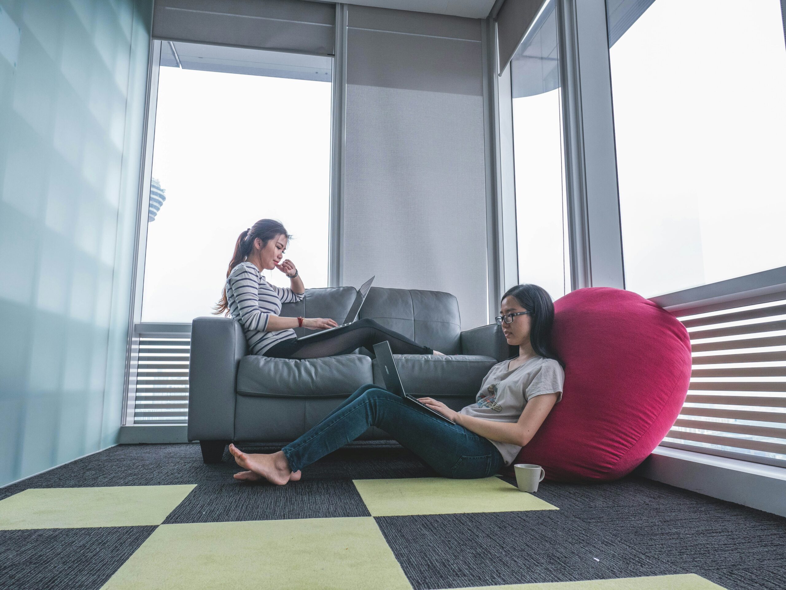 A young woman lounged on a couch with a laptop and next to her is another woman on the floor with a laptop, leaning against a large pillow. 